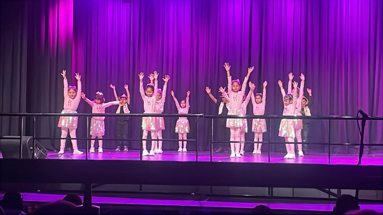 A groups of students are dressed in costume for their dance performance. They are mid routine and have their hands above their heads.