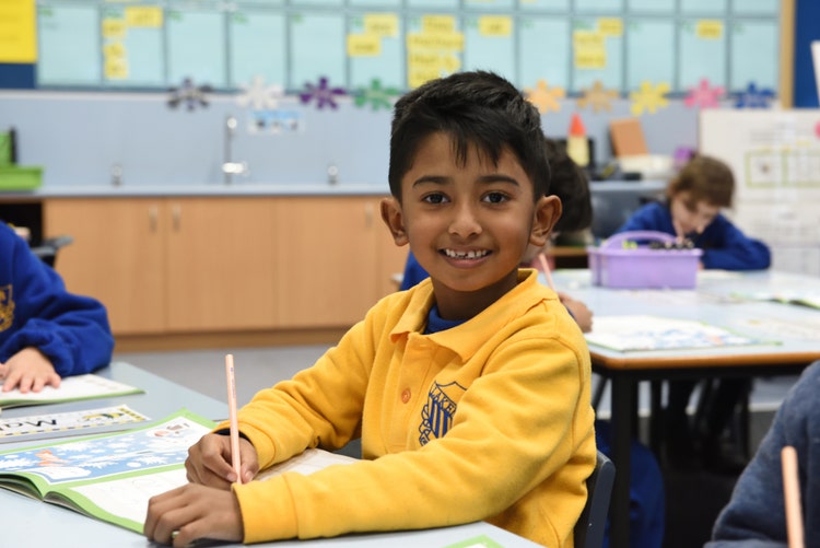 A boy is sitting at his desk, holding a pencil and smiling.