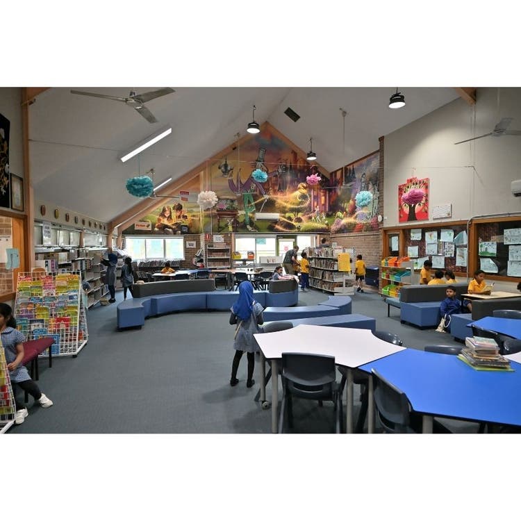 This is a photo of the school library. There are groups of tables, some children standing around, books on shelves and a colourful mural on the wall at the back.