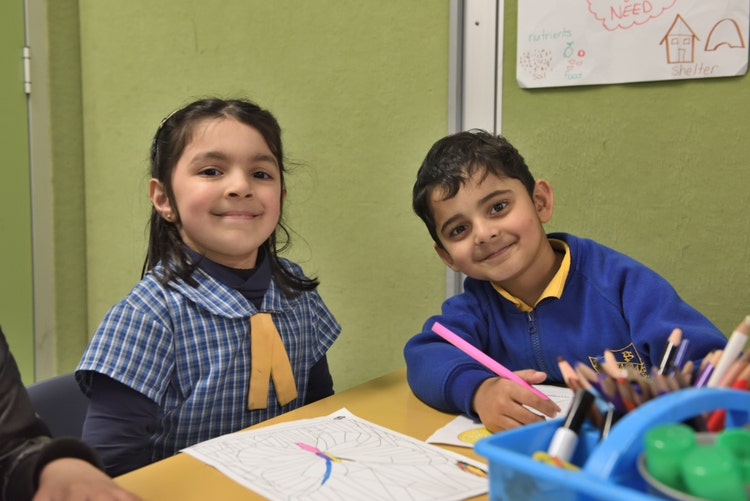 A girl and boy are sitting at a desk and are smiling. The boy is holding a pencil.