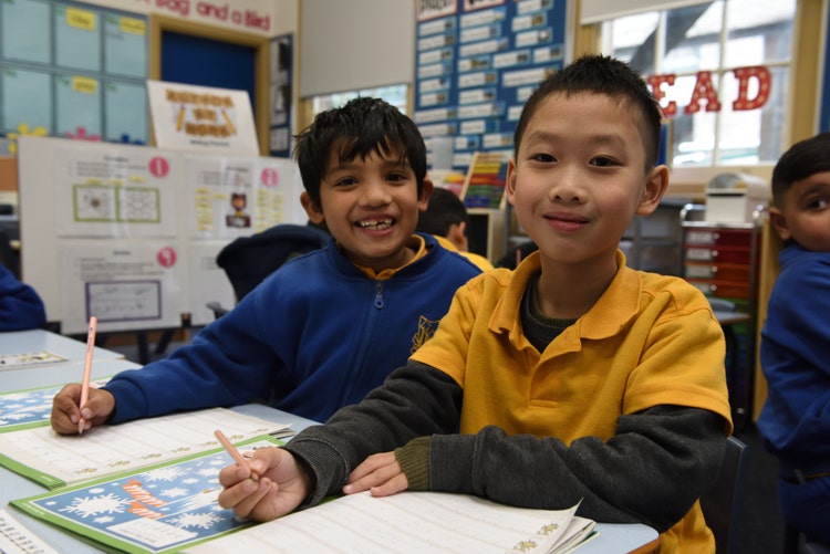 Two boys are sitting at their desks and smiling. They are holding pencils and have work on their desks.