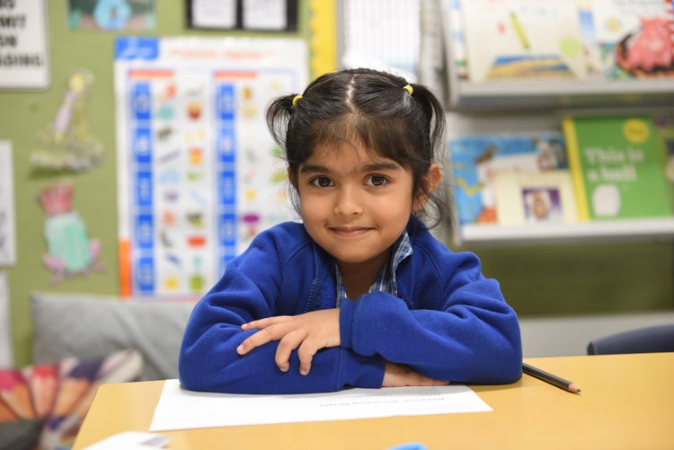 One girls sits and her desk and is smiling. Her arms are folder and she has her arms on a piece of paper.