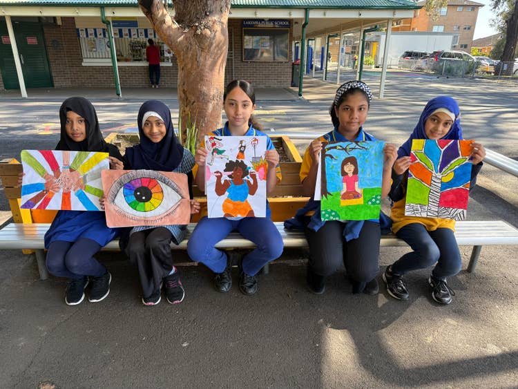 This shows five female students sitting on a bench in the playground holding up artworks they have created.