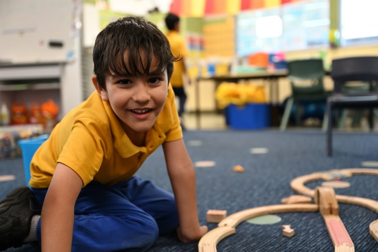 This is a photo of a boy playing on the floor with a train set and smiling.